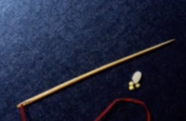 Image 67: A long brown needle with red thread coming out of the eye, along with a white bead and gold beads is seen on a blue surface